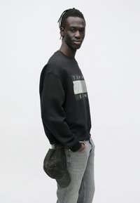 Sudadera negra con un gráfico rectangular en el pecho, vaqueros grises y una gorra de béisbol oscura metida en la cintura.