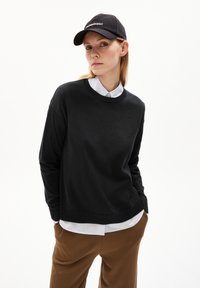 Schwarzer Rundhalsausschnitt-Pullover über einem weißen Kragenhemd, getragen mit braunen Hosen. Das Model trägt eine schwarze Mütze mit dem Wort "PERMANENT."