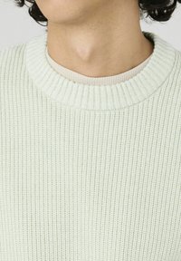 Pull en mailles côtelées vert clair avec un col rond, doté d'un motif tricoté texturé et d'un sous-vêtement beige superposé visible au niveau du col.