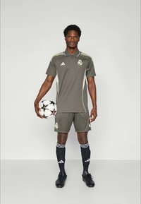 Mandlig fodboldspiller i olivengrønt Real Madrid-sæt, der holder en hvid og sort UEFA Champions League-bold, stående mod en ensfarvet baggrund.