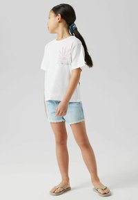 Camiseta blanca de manga corta con un gráfico de sol rosa, combinada con unos pantalones cortos de denim azul claro deshilachados y chanclas beige.