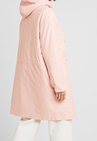 Jasnoróżowy długi płaszcz z kapturem, z zapięciem na zamek, bocznymi kieszeniami i lekko luźnym krojem. Gładka tkanina o matowym wykończeniu.