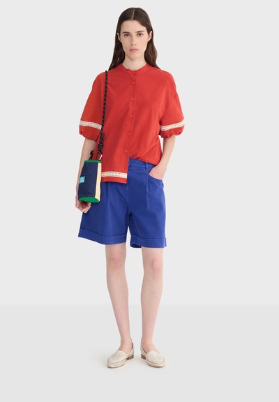 Femme portant un chemisier rouge boutonné avec des manches bouffantes et un short bleu jusqu'aux genoux, tenant un petit sac bandoulière bleu marine et beige.