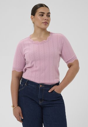 Femme portant un haut en maille à manches courtes rose clair avec bordure en dentelle, assorti à un jean bleu foncé, posant avec une main dans la poche.