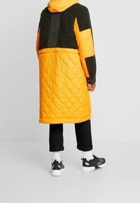 Vadderad lång jacka med orange nederdel och mörkgrön överdel, med en huva och kontrasterande tygtexturer. Bärs med svarta byxor och sneakers.