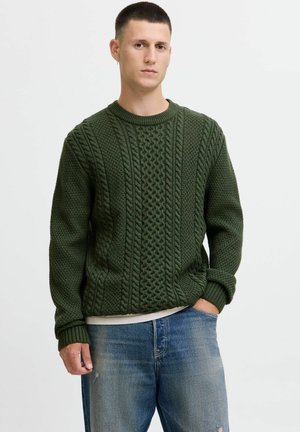 Ein Mann trägt einen dunkelgrünen Grobstrickpullover und eine blaue Jeans. Er steht mit einer Hand in seiner Tasche vor einem schlichten Hintergrund.