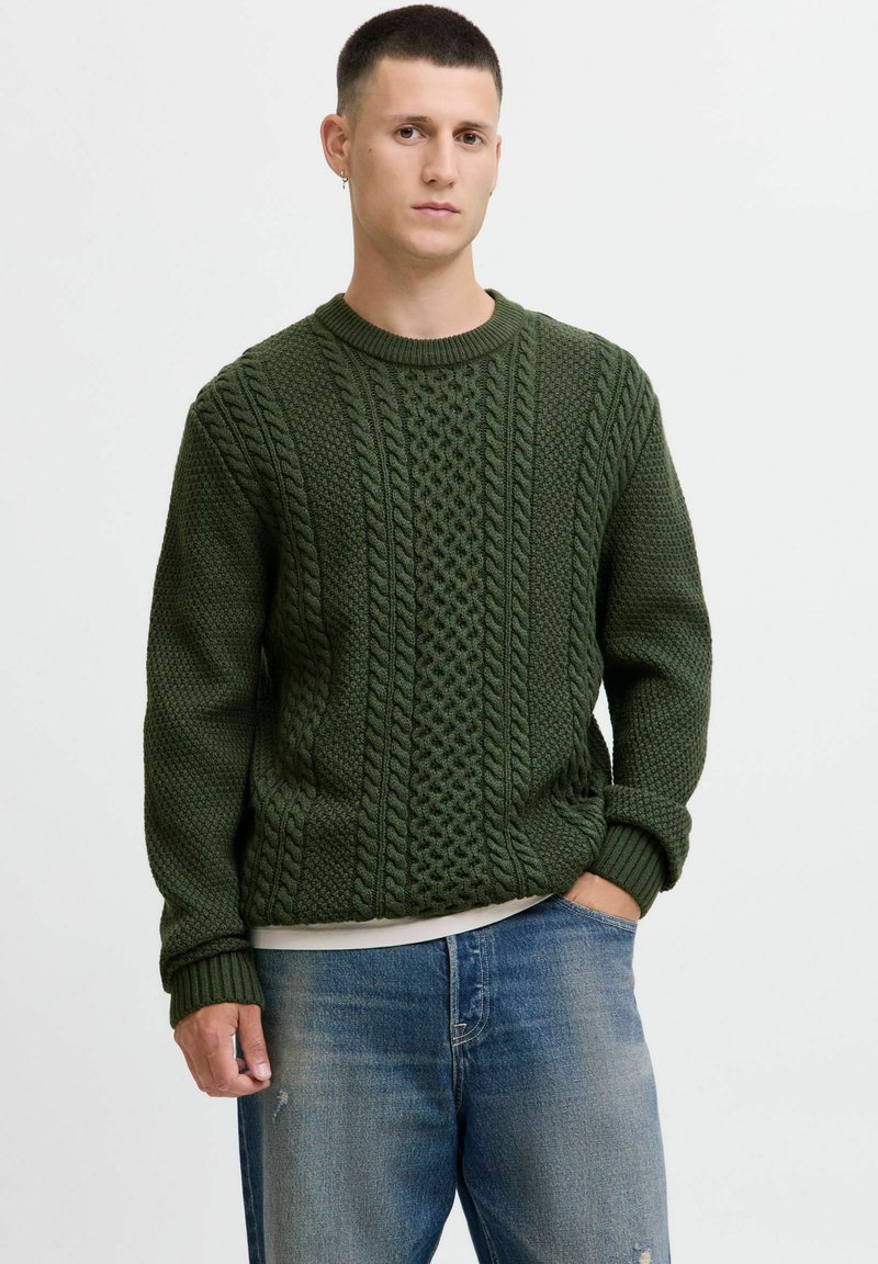 Mand iført en mørkegrøn kabelstrik sweater og blå jeans, står med den ene hånd i lommen mod en ensfarvet baggrund.