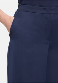 Pantalon bleu marine en tissu lisse et texturé, avec des poches latérales et une coupe ajustée avec un léger brillant.