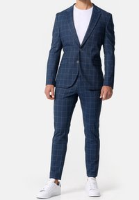 Traje a cuadros azul marino con diseño entallado, con solapa con muesca y cierre de un solo botón. Combinado con zapatillas blancas.