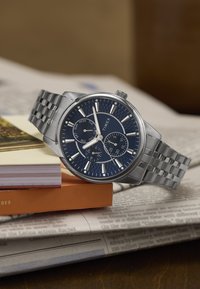 Reloj de acero inoxidable plateado con un dial redondo de color azul marino, marcadores de hora plateados y una pulsera; cuenta con subdiales de fecha y día.