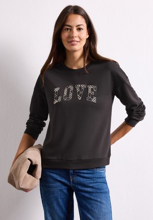 Femme portant un sweat-shirt noir avec le texte "LOVE", tenant une veste beige pliée, debout devant un fond clair uni.