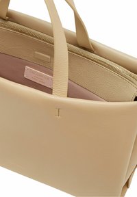 Beige lædertaske med en glat tekstur, to håndtag og en pink stofforing med en logoetiket. Lynlås lukning på toppen.