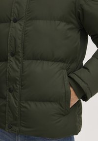 Green padded jacket featuring a quilted design, black snap buttons, and side pockets. Made of smooth, shiny material with a textured finish.