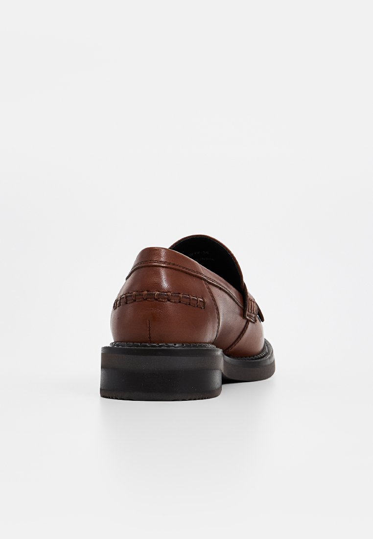 Zapato marrón de cuero tipo slip-on con un patrón trenzado en el tacón y una suela de goma negra. El diseño es limpio con acentos mínimos.