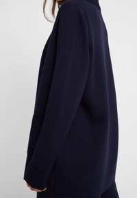 Cardigan azul marino de ajuste relajado, con textura acanalada, mangas largas y bolsillos laterales. El dobladillo presenta un sutil detalle de costura roja.