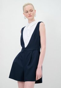 Mono sin mangas azul marino con detalle de camisa blanca con cuello, que presenta un escote en forma de V, pantalones cortos con pleat y una textura de tela suave.