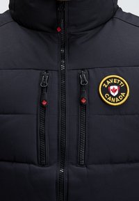 Giacca nera imbottita con cerniera centrale, due cerniere verticali sul petto con tiranti a forma di foglia d'acero rossa e patch del logo Zavetti Canada bordato di giallo.