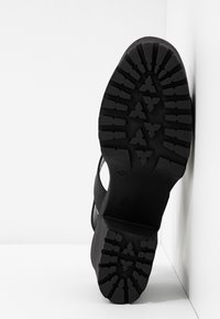 Semelle noire d'une chaussure à talon avec des motifs de bande de roulement profonds, appuyée contre un coin blanc entre mur et sol.