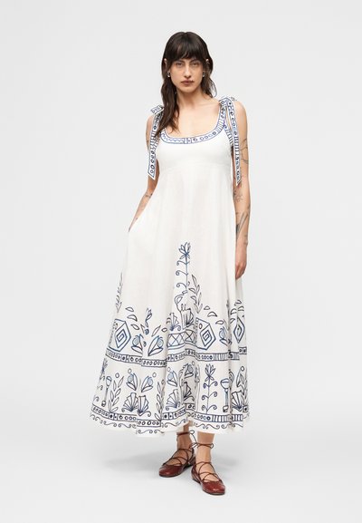 Γυναίκα που φοράει λευκό αμάνικο μάξι φόρεμα με μπλε λουλουδάτα και γεωμετρικά σχέδια, δεμένα τιράντες στους ώμους και καφέ δερμάτινα επίπεδα παπούτσια με κορδόνια.