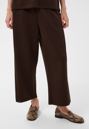 Persona che indossa pantaloni larghi marrone scuro con dettagli a bottoni sull'orlo e mocassini slip-on con fantasia effetto pelle di serpente su pavimento bianco.