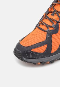 Gros plan d'une chaussure de running orange avec mesh noir et superpositions synthétiques, coutures détaillées et lacets noirs sur fond blanc.