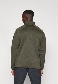 Grüner Fleece-Pullover mit hohem Kragen, langen Ärmeln und strukturiertem Finish. Verfügt über eine Seitentasche und ein dezentes Branding-Detail auf der Schulter.