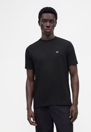 Giovane uomo dalla pelle scura che indossa una semplice t-shirt nera a maniche corte con scollo rotondo e pantaloni scuri su sfondo bianco.
