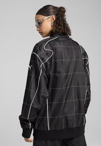 Jachetă bomber din satin negru cu model în carouri albe, cu manșete și tiv ribbed. Prezintă logo pe mânecă și detalii de cusătură în contrast.
