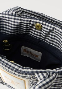 Εσωτερικό υφασμάτινης τσάντας σε ναυτικό μπλε και λευκό καρό, με μπρούτζινο κουμπί πατιλέτας, τσέπη με φερμουάρ και ετικέτα με την επωνυμία Barbour.