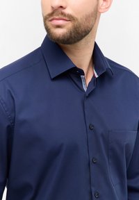 Camicia button-up blu navy realizzata in tessuto morbido, con una piccola tasca sul petto e un motivo a righe all'interno del colletto.