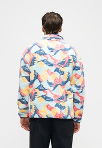 Άνδρας που φορά πολύχρωμο φλις μπουφάν με αφηρημένα μοτίβα βουνών και ήλιου, όρθιος, γυρισμένος με την πλάτη προς φόντο απλού ανοιχτού χρώματος.