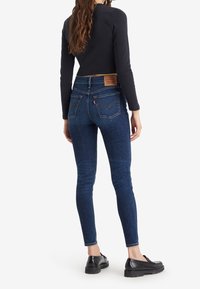 Mujer con cabello largo castaño, vestida con jeans ajustados oscuros, una blusa negra de manga larga y mocasines negros, de pie de espaldas sobre un fondo blanco.