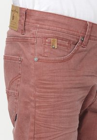 Gros plan d'un pantalon en jean rouge délavé avec poches avant et latérales visibles, rivets métalliques et écusson en cuir de la marque sur la ceinture.