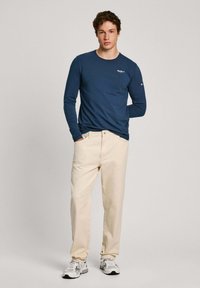 Camisa de manga larga azul marino con cuello redondo, pantalones beiges y zapatillas grises. La camisa cuenta con un pequeño logo en el pecho y un diseño sencillo.