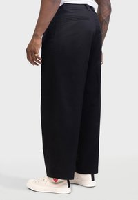 Zwarte wijde broekspijpen gemaakt van stof met een gladde textuur. Voorzien van zijzakken, een achterzak en een omslagzoom boven witte schoenen.