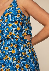 Květinové šaty s modrým pozadím a žlutými a oranžovými květy, s volánkovým zakončením a výřezem v pase. Vyrobeno z lehkého materiálu.