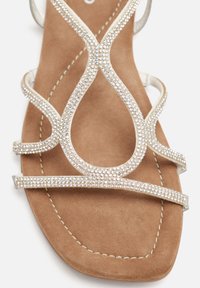 Sandal piatta con fondo in suede color tan, con cinturini in strass argentati brillanti disposti a incrocio e un accent gemma rotonda al centro.