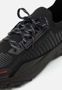 Černá sportovní bota s síťovaným svrškem, texturovanými gumovými akcenty, plochými tkaničkami a červeným logem. Má drsnou podrážku pro trakci.