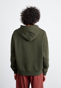 Olive green sweatshirt with a hood, soft texture, ribbed cuffs and hem, loose fit. Worn with burgundy pants, viewed from the back.