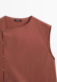 Ärmlös topp i terrakottafärgad tyg med en mjuk yta. Har rund halsringning och sidoknäppning för stängning. Varumärkesetikett synlig.