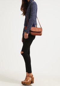 Sac bandoulière en cuir marron, pull côtelé bleu marine, chemise à motifs, jean skinny noir déchiré, et sandales à talons épais marron.