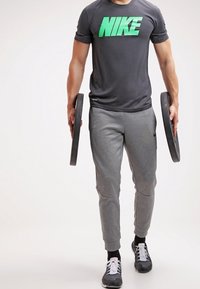 Szary t-shirt Nike z zielonym logo, połączony z szarymi dresami. Osoba trzyma dwa ciężarki, ma na sobie czarne sneakersy z białymi akcentami.