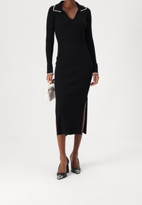 Svart ribbad stickad klänning med V-ringning, utsmyckad krage och långa ärmar. Har sidoklyftor och är kombinerad med svarta klackar och en krämfärgad handväska.