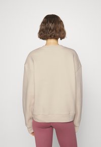 Personne avec les cheveux bruns courts portant un sweat-shirt beige à manches longues et des leggings mauves, debout face à un fond blanc uni.
