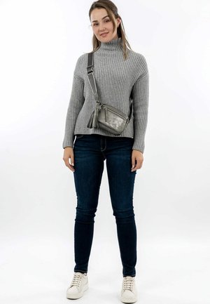 Young woman wearing a gray turtleneck sweater, dark jeans, white sneakers, and a crossbody metallic gray bag, standing against a white background.