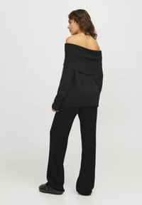 Zwarte off-shoulder gebreide trui met lange mouwen, gecombineerd met zwarte wijde broek en instap schoenen, gezien vanaf de achterkant.
