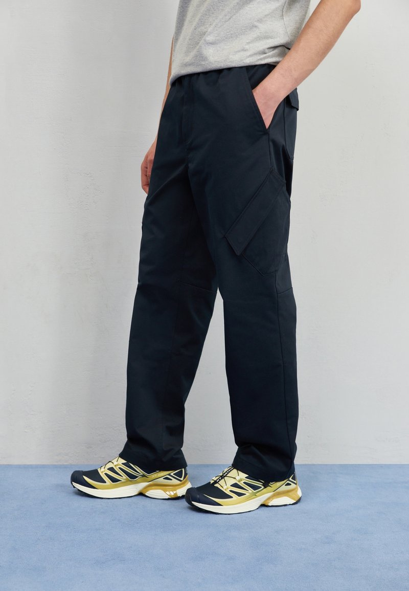 Personne portant un pantalon cargo noir avec des poches et des chaussures de course jaunes et noires, debout sur un tapis bleu contre un mur uni.