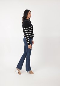 Striped black and white sweater, dopasowany, z kołnierzykiem. Dżinsy w fasonie dzwonów, ciemny odcień. Przezroczyste szpilki. Neutralne tło.