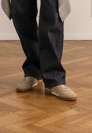 Persona con jeans blu scuro larghi e scarpe da ginnastica in pelle scamosciata beige che stanno in piedi su un pavimento in parquet di legno.