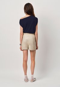 Marinel blauw off-shoulder top gecombineerd met beige leren shorts met metalen studs langs de zoom. Model staat met de rug naar de camera.